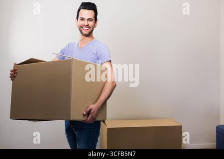Homme adulte moyen debout tenant une grande boîte en carton ouverte près de la boîte scellée dans une pièce vide Banque D'Images