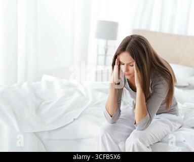 Femme à la fin des années 20 assise sur le lit pressant les temples dans la chambre à coucher près de la lampe moderne, espace de copie Banque D'Images