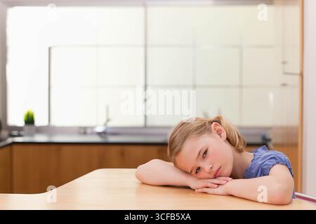 Fille d'âge scolaire appuyée sur la table dans la cuisine près de l'évier en acier inoxydable et de l'usine, espace de copie Banque D'Images
