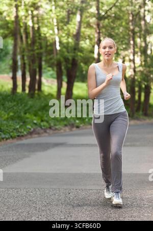 Coureuse femme jogging sur une route forestière pavée dans un débardeur gris et leggings entouré d'arbres Banque D'Images