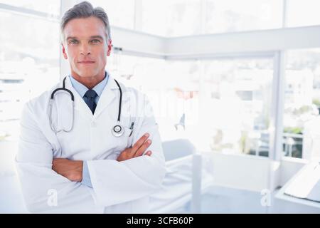 Médecin masculin mature debout avec les bras pliés dans la clinique médicale avec stéthoscope, blouse de laboratoire Banque D'Images