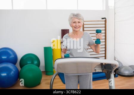 Femme senior debout sur le tapis roulant soulevant des haltères sarcelle au niveau des épaules au gymnase avec des ballons d'exercice Banque D'Images