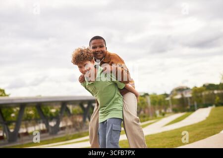 Garçon caucasien donnant piggyback ride à Black Boy en plein air, à la fois souriant et regardant la caméra, des amis appréciant un moment ludique ensemble dans le cadre du parc Banque D'Images
