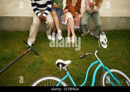 Groupe d'adolescents assis sur un rebord en béton mangeant des collations, jambes visibles, garçons et filles multiethniques se relaxant à l'extérieur avec vélo, scooter et planche à roulettes sur l'herbe au premier plan Banque D'Images
