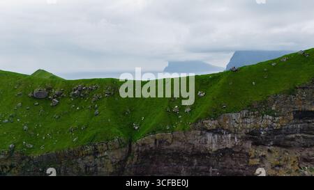 Les falaises vertes roulantes surplombent l'océan serein des îles Féroé, avec des montagnes lointaines entourées de brume. Un paysage paisible et dramatique showcas Banque D'Images