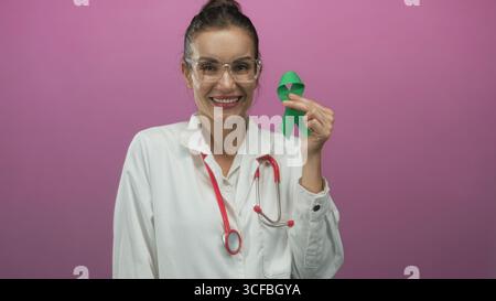 Jeune femme hispanique souriante médecin tenant un ruban vert dans un studio rose ; espoir de sensibilisation à la santé mentale. Banque D'Images