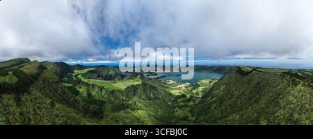 Vue aérienne des collines verdoyantes en cascade vers la sereine Lagoa das Sete Cidades, reflétant le ciel sous une couverture de nuages, Açores, Portugal. Banque D'Images