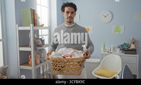 Jeune homme hispanique tient un panier à linge dans un intérieur confortable rempli de jouets pour enfants et la lumière douce du jour qui traverse les fenêtres créant un serein Banque D'Images
