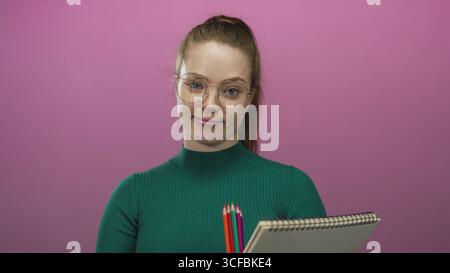 Femme portant des lunettes tient le carnet de croquis et les crayons de couleur avec le geste sourire dans le studio rose vif ; créativité curiosité inspiration doute. Banque D'Images