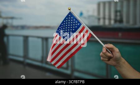 Homme tenant le drapeau américain dans un port de bord de mer, soulignant le patriotisme et l'atmosphère maritime, avec un fond de bateau flou créant un cadre côtier. Banque D'Images