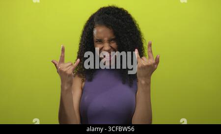 Jeune femme aux cheveux bouclés et aux bras levés montre un geste de cornes de roche dans un cadre de studio jaune ; ambiance amusante. Banque D'Images