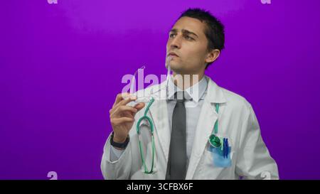 Jeune médecin masculin hispanique en uniforme avec stéthoscope contemplant au-dessus des lunettes sur fond violet isolé mettant en valeur l'accent professionnel et Banque D'Images