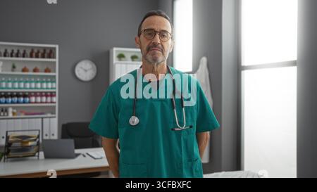 Médecin masculin hispanique dans des gommages verts avec stéthoscope debout en toute confiance dans un bureau d'hôpital avec des documents médicaux et de l'équipement en arrière-plan Banque D'Images