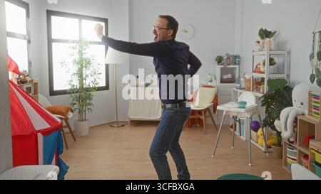 Homme dansant joyeusement dans une crèche colorée remplie de jouets et de meubles, mettant en valeur une atmosphère intérieure heureuse et animée. Banque D'Images