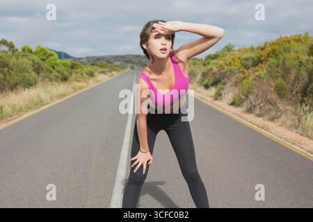 Femme portant un soutien-gorge de sport rose et des leggings noirs penchés vers l'avant protégeant les yeux sur la route pavée rurale Banque D'Images
