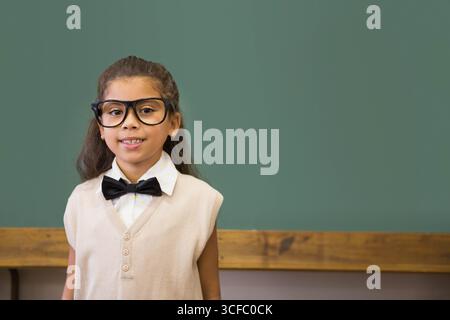 Enfant hispanique debout au plateau de craie du tableau noir dans la salle de classe portant des lunettes noeud papillon, espace de copie Banque D'Images
