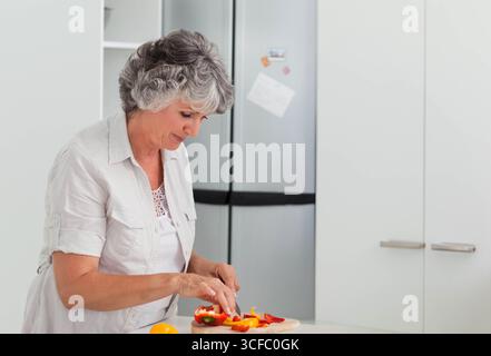 Femme senior tranchant des poivrons jaunes et rouges sur une planche à découper dans une cuisine blanche, espace de copie Banque D'Images