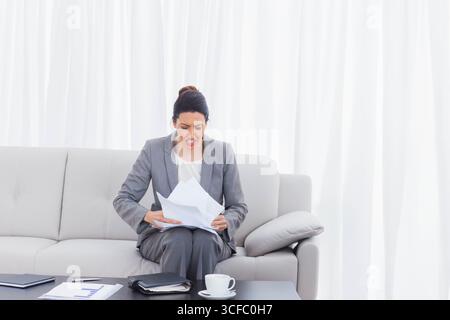 Femme d'affaires portant costume assis sur le canapé dans le salon tenant des papiers et grimacant près de la tasse de café Banque D'Images