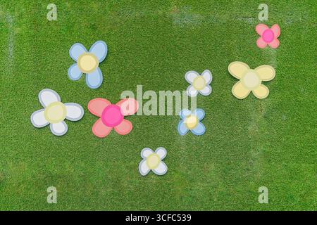 huit fleurs de papier colorées dispersées sur l'herbe verte. Des fleurs de couleurs vives en bleu, rose, jaune et blanc contrastent avec le fond naturel Banque D'Images