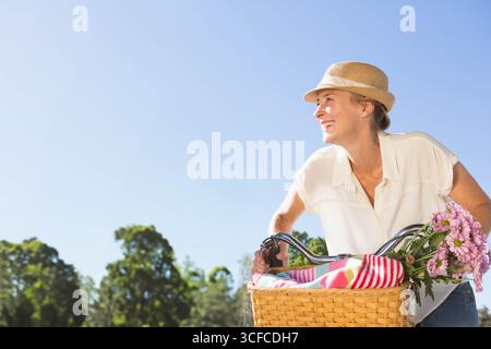La femme fait du vélo sur le chemin ensoleillé du parc, avec un panier en osier tenant une couverture rayée et des marguerites Banque D'Images