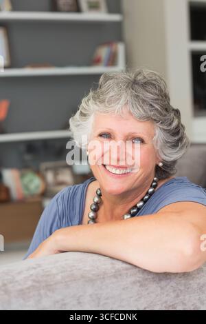 Femme senior appuyée sur le dossier gris du canapé à la maison, sourire clignotant avec des livres sur l'étagère blanche Banque D'Images