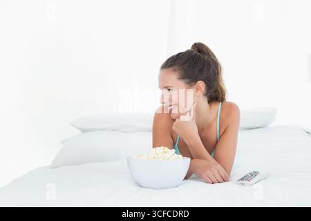 Femme s'inclinant sur un couvre-lit blanc dans un débardeur turquoise, atteignant un bol de pop-corn près de la télécommande Banque D'Images