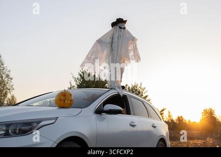 Enfant méconnaissable en couverture blanche, chapeau, lunettes noires se tient sur le toit de la voiture blanche au coucher du soleil. Costumes fantômes d'Halloween. Farces de vacances. Trick ou tr Banque D'Images