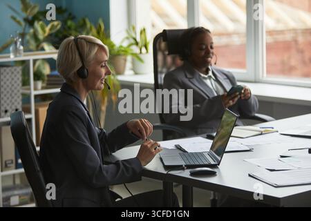 Femme caucasienne d'âge moyen portant un casque travaillant sur un ordinateur portable au bureau tandis que jeune femme adulte noire en costume d'affaires assis à proximité à l'aide d'un smartphone Banque D'Images