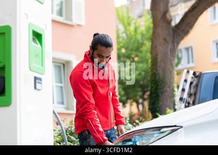 Homme en veste rouge chargeant la voiture électrique à la station dans le quartier résidentiel. Allemagne Banque D'Images