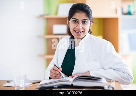 Femme en manteau blanc assise à un bureau avec un carnet, souriant chaleureusement à la caméra. Allemagne Banque D'Images
