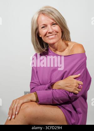 Femme souriante en haut violet assise, bras croisés, cheveux blonds courts, fond neutre. Italie Banque D'Images