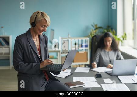 Femme caucasienne d'âge moyen portant un casque à l'aide d'un ordinateur portable tout en se tenant debout dans un bureau moderne, jeune femme biraciale travaillant au bureau avec des documents et un ordinateur en arrière-plan Banque D'Images