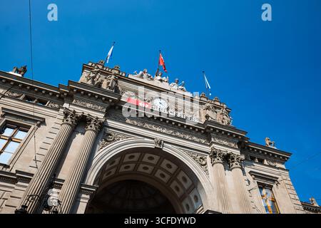 Zurich, Suisse, 19 août 2025. Façade d'entrée du bâtiment de la gare centrale de Zurich HB. Regardant vers le haut shot, jour d'été ensoleillé, grande horloge et drapeau, pas de poo Banque D'Images