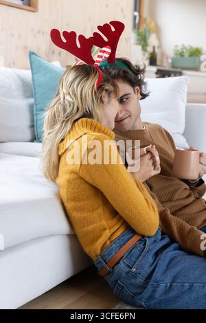 Couple tenant des tasses portant des bandeaux en bois rouge appuyé contre le canapé avec oreiller sarcelle et regardant Banque D'Images