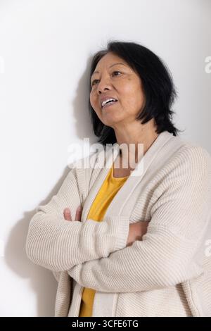 Femme asiatique senior appuyée contre le mur blanc dans le studio portant chemise moutarde et cardigan beige Banque D'Images