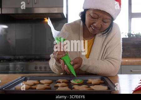 Femme asiatique senior penchée sur le comptoir de cuisine, tapissant du glaçage vert sur les biscuits, espace de copie Banque D'Images