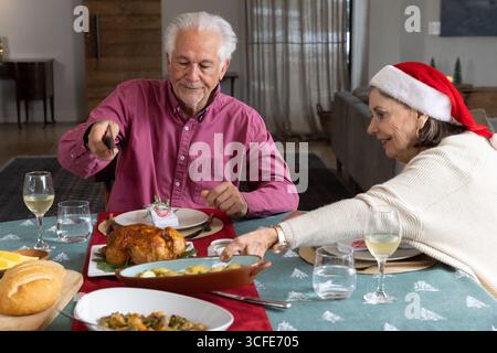 Couple senior portant des vêtements de fête sculptant la dinde et passant cocotte à la table à manger Banque D'Images