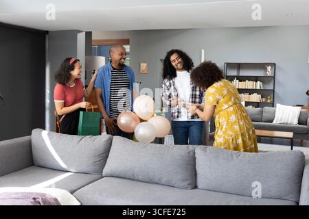 Divers amis se réunissant dans le salon au canapé, remettant le cadeau emballé, tenant des ballons pastel Banque D'Images