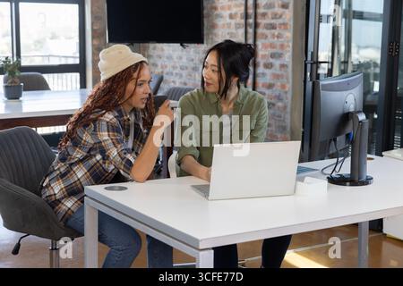Diverses collègues féminines collaborant sur ordinateur portable et moniteur au bureau blanc dans un bureau ouvert Banque D'Images