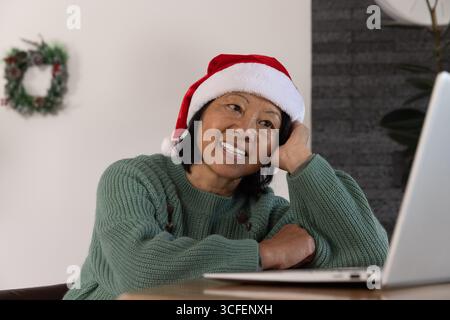 Femme asiatique senior assise à table à la maison portant chapeau de Père Noël à l'aide d'un ordinateur portable avec couronne, plante Banque D'Images