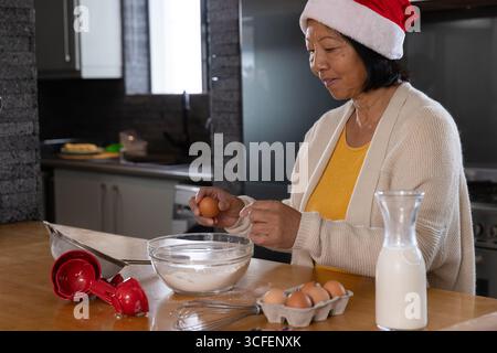 Femme asiatique senior portant chapeau de Père Noël craquant l'oeuf dans le bol sur le comptoir dans la cuisine, copiez l'espace Banque D'Images