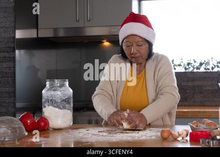 Femme asiatique senior portant chapeau de Père Noël rouge pétrissant la pâte sur le comptoir de cuisine avec pot de farine Banque D'Images