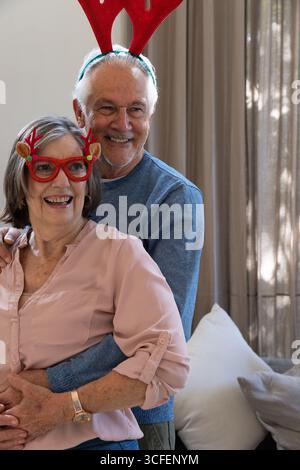 Couple senior portant des bandeaux en bois serrant et souriant à côté des coussins de canapé dans le salon Banque D'Images