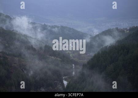 Une vallée paisible s'étend entre des collines couvertes de forêts, un ruisseau étroit serpentant son chemin à travers le paysage. La brume s'élève des arbres Banque D'Images