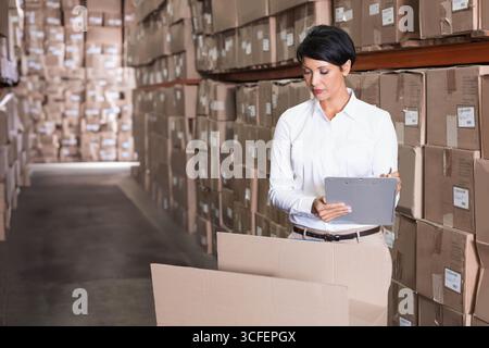 Femme vérifiant l'inventaire dans l'allée de l'entrepôt avec presse-papiers gris et boîte en carton ouverte, espace de copie Banque D'Images