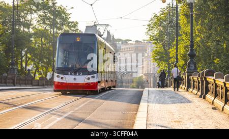 Un tramway longe le pont de la Légion à Prague, en République tchèque. La scène capture la lumière chaude de la soirée, mettant en valeur les arbres environnants et l'architecture historique. Banque D'Images