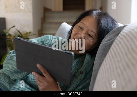 Femme asiatique senior s'inclinant sur un canapé rembourré contre un oreiller dans le salon tenant la tablette Banque D'Images