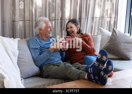 Couple senior souriant tout en tenant des tasses rouges sur le canapé de couleur claire dans le salon confortable Banque D'Images
