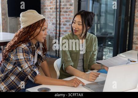 Collaboratrices féminines diverses collaborant au bureau dans le bureau avec un ordinateur portable, des documents et un distributeur de ruban adhésif Banque D'Images