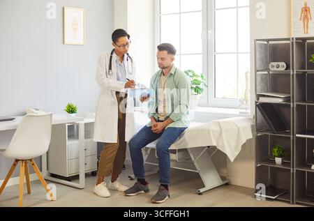 Patient masculin assis sur la table d'examen, médecin féminin tenant une presse-papiers, expliquant les résultats du test Banque D'Images